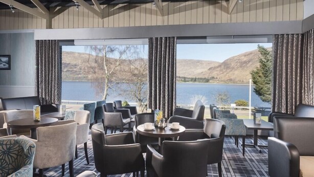 A hotel room with a large window offering a view of a lake and mountains. There are multiple seating areas with tables and chairs. - Caledonian Hotel (Fort William)
