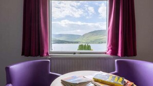 A room with a window view of a lake and hills, purple chairs, and a table with books. - Caledonian Hotel (Fort William)