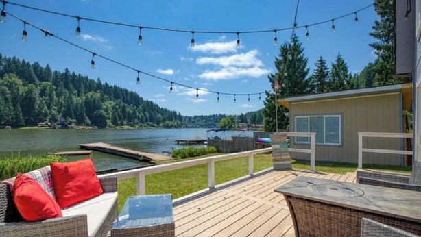 A deck with wicker furniture and a small table, overlooking a lake with a dock and a building in the background.