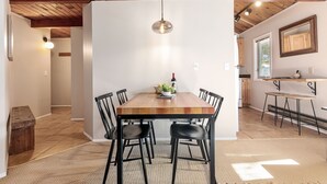 A dining area with a wooden table and black chairs, a hanging light fixture, and a kitchen in the background.