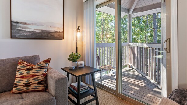A cozy living room with a sofa, a small table with a lamp, and a view of a wooden deck through glass doors.