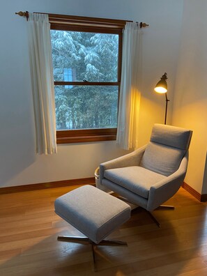 Una habitación con un ventanal grande, una silla, un reposapiés y una lámpara de pie.