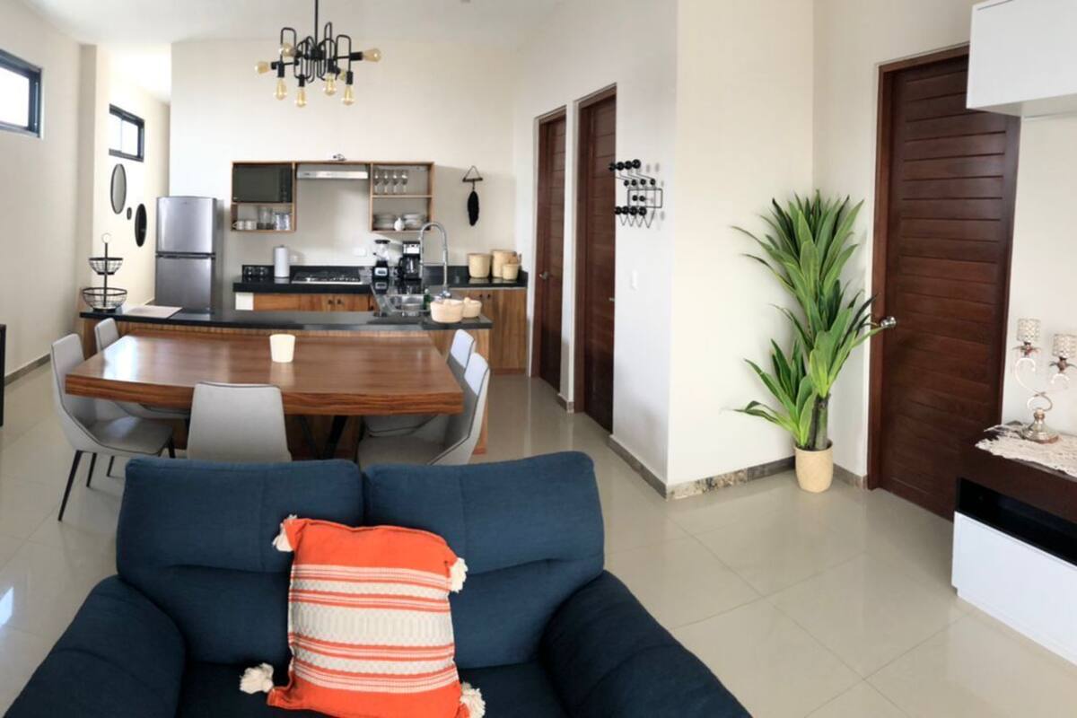 A modern living room with a dark blue sofa, a dining table with chairs, and a kitchen area with a refrigerator and stove.