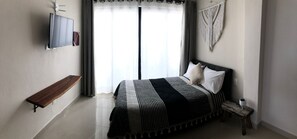 A bedroom with a bed, a TV, a bench, and a macrame wall hanging. - Casa Luz Downtown Apartments Tulum (Tulum)