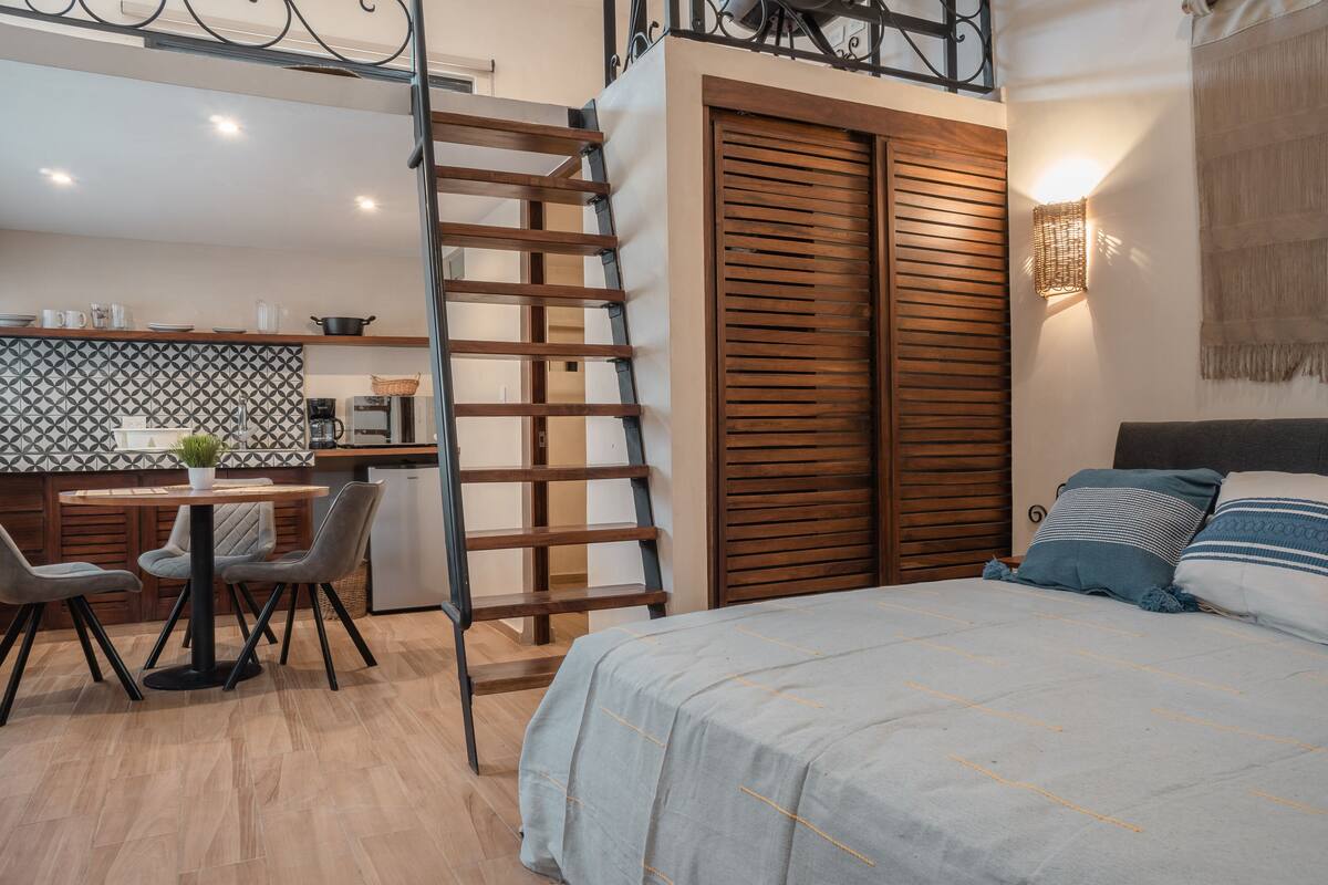 A bedroom with a bed, bedside tables, and a wardrobe. There is a kitchen area with a counter, chairs, and a ladder.