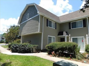 A two-story residential building with a gabled roof, a front porch, and a small front yard with bushes and a pathway.