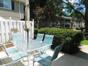 A patio with a glass table and chairs, surrounded by a white fence and greenery.