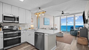 Une cuisine moderne avec des armoires blanches, des appareils en acier inoxydable et un grand îlot central. Un balcon offre une vue sur l’océan.