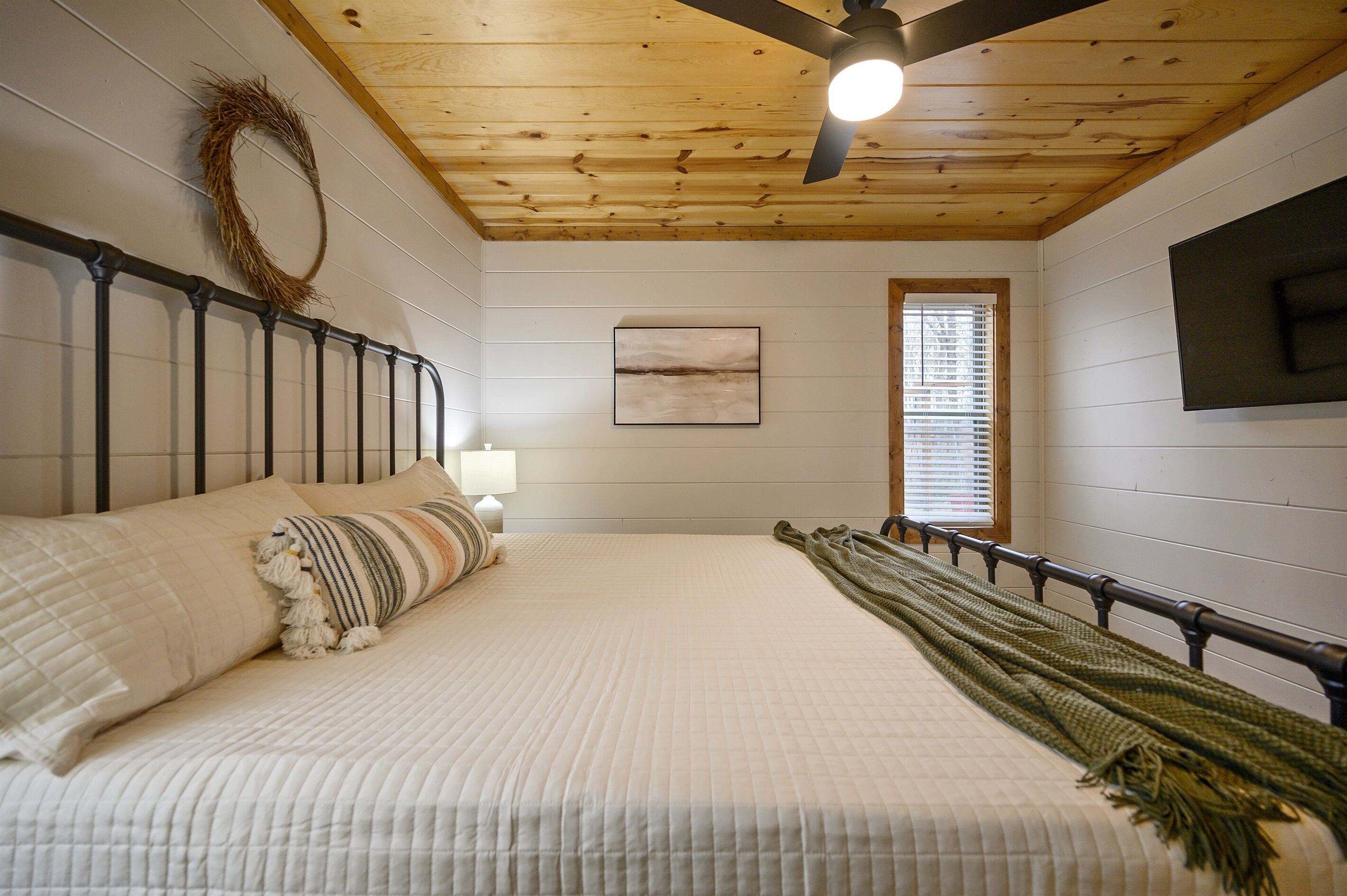 A bedroom with a bed, a ceiling fan, a window with blinds, and a wall-mounted TV.