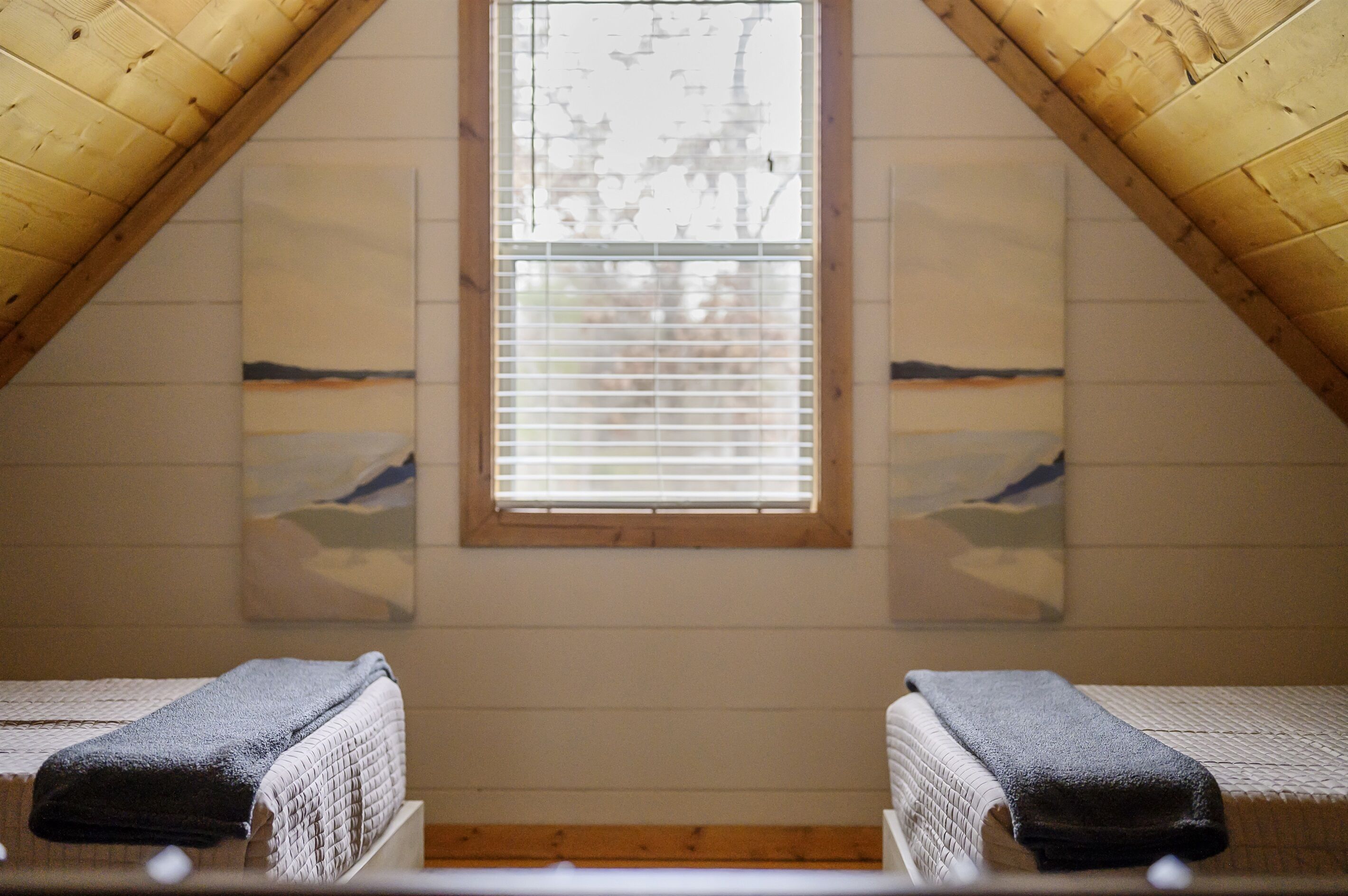 A room with two beds, a wooden ceiling, and a window with blinds.