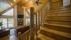 A wooden staircase with a ceiling fan, a stone fireplace, and a leather sofa in a spacious living area with large windows. - ❤️Heart of Hochatown | Modern Fam Friendly Cabin, Fun Games, Hottub, Firepit ✨ (Broken Bow)