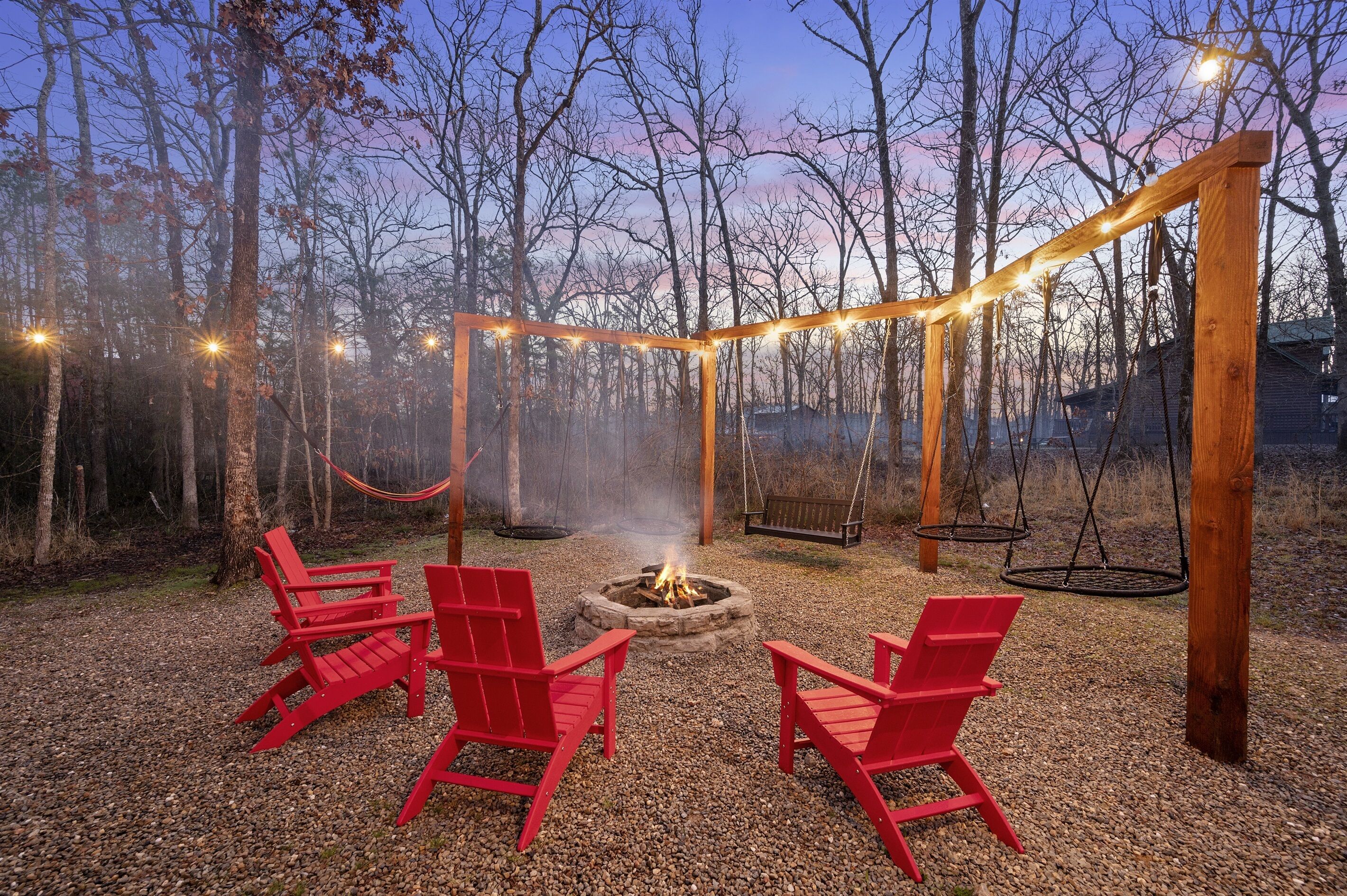 Una pérgola de madera con luces colgantes, un brasero y sillas Adirondack rojas en un área boscosa.