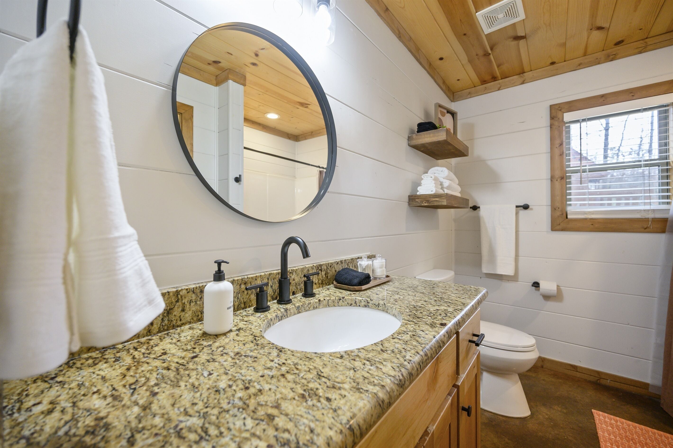 Un baño con encimera de granito, un lavabo blanco, un espejo, inodoro, estanterías de madera y una ventana.