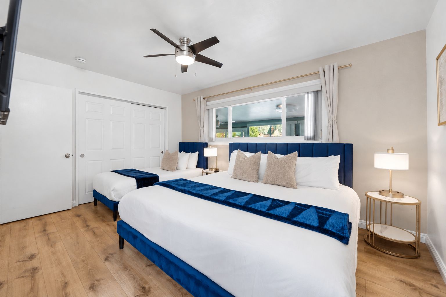 A hotel room with two beds, a ceiling fan, a window with curtains, and a bedside table with a lamp.