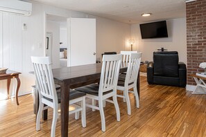 A dining area with a wooden table and white chairs, a black leather sofa, and a TV mounted on a brick wall.