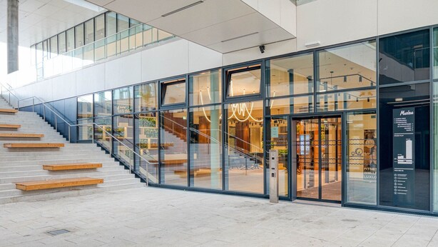 Un edificio moderno con fachada de vidrio, una escalera que conduce a un nivel superior y un cartel que dice 'Marina' a la derecha.
