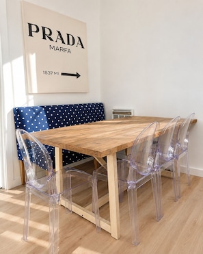 Zona pranzo con tavolo in legno, sedie in plastica trasparente, una panca a pois e un cartello appeso al muro con la scritta 'PRADA MARFA 1837 MI'.