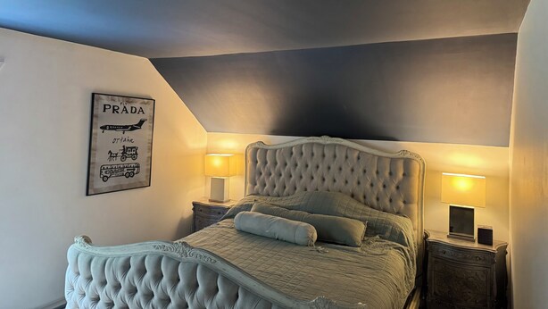 Een slaapkamer met een gestapeld hoofdeinde, twee nachtkastjes met lampen, een ingelijst poster aan de muur en een bed met een gewatteerde spreideken.