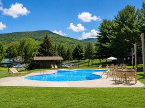 A swimming pool with lounge chairs and a wooden building, surrounded by green grass and trees, with a mountain in the background. - Jiminy Peak Resort Townhome, Sleeps 10, Pool, Hot Tubs, Sauna & Deck (Hancock)