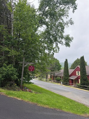 A rural road with a wooden signpost, a red building, and a grassy area with flowers. - On Jiminy Peak Resort - Sleeps 9-10 - Pool, Hot Tub & Sauna (Hancock)
