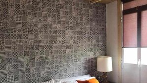 A bed with white bedding and orange pillows, a patterned wall, and a wooden ceiling.