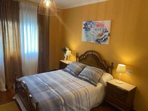 A bedroom with a bed, two bedside tables, a hanging light fixture, a painting on the wall, and a window with curtains. - VOLVORETA APARTMENT in the center of O Grove.             (Grove, O (Casco Urbano))