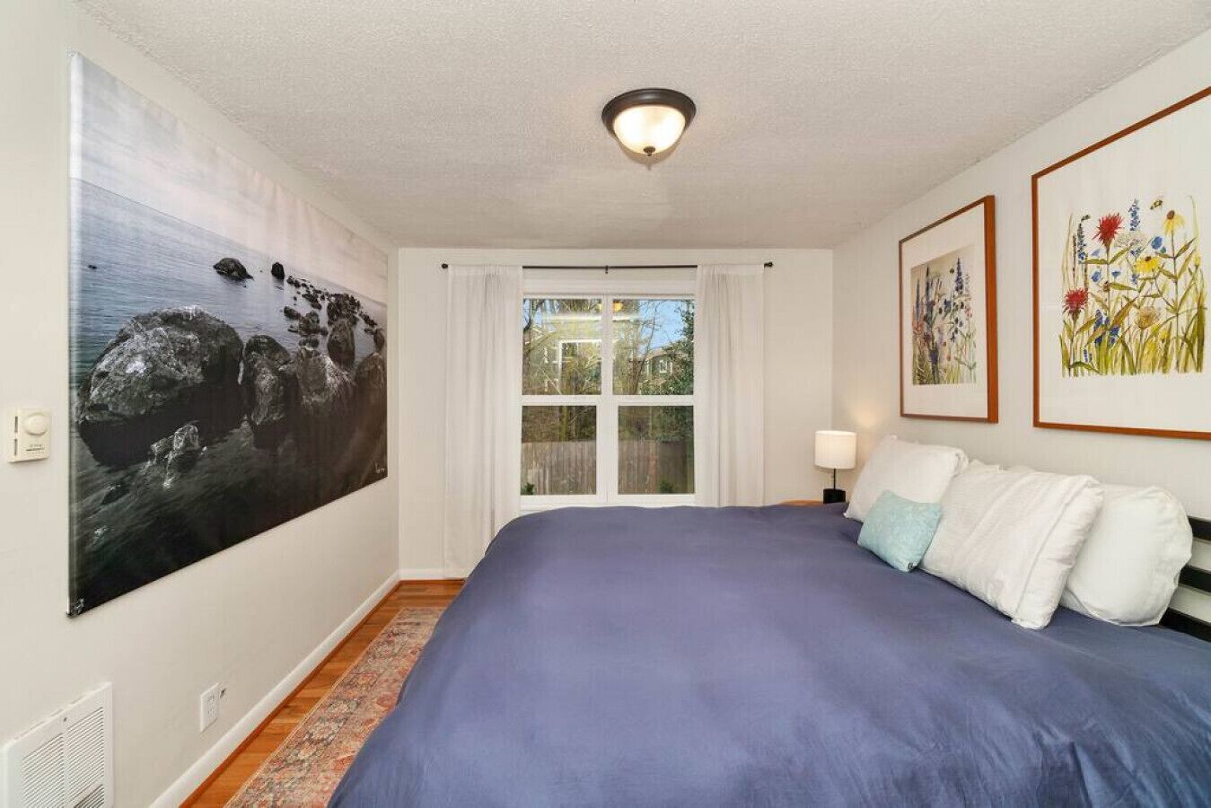 A bedroom with a large bed, a window, a ceiling light, and framed artwork on the walls.