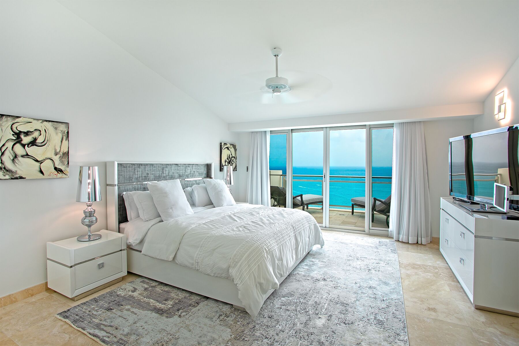 A bedroom with a bed, a nightstand, a lamp, a TV, and a view of the ocean through large sliding doors.