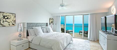 A bedroom with a bed, a nightstand, a lamp, a TV, and a view of the ocean through large sliding doors.