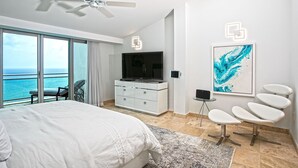 A modern hotel room with a large bed, a ceiling fan, a TV, a desk, a chair, and a view of the ocean. - The Cove | Beach Front - Located in  Exquisite Simpson Bay with House Cleaning Included (Lowlands)