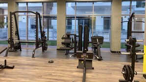 Een sportschool met diverse trainingsapparaten, grote ramen en uitzicht op een witte schutting.