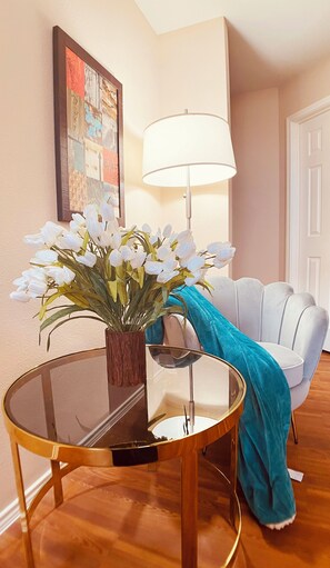 Una mesa redonda de vidrio con un florero de flores blancas, una lámpara y una manta azul sobre una silla blanca.
