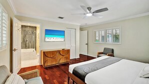 A bedroom with a bed, a TV, a ceiling fan, a shower, and a window with blinds.