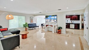 A spacious living room with a sofa, coffee table, and fireplace. There is a dining area with a table and chairs, and a kitchen with dark cabinets.