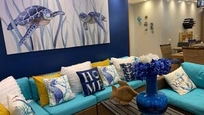 Un salon avec un canapé de couleur turquoise, des coussins décoratifs, un vase bleu et un tableau représentant des tortues de mer.