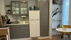 Een moderne keuken met grijze kastjes, een witte koelkast en een magnetron. Er is een eethoek met een tafel en stoelen, en een klein plankje met decoratieve spullen.
