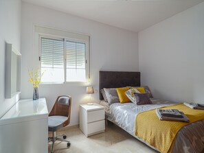 Ein Schlafzimmer mit Bett, Nachttisch, Stuhl, Lampe und einem Fenster mit Jalousien.
