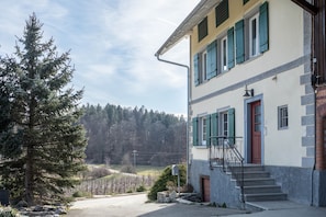 A two-story building with a red door, a staircase, and a view of a forested area. - Apartment 'Am Hopfengarten' with Mountain View, Garden & Wi-Fi (Tettnang)