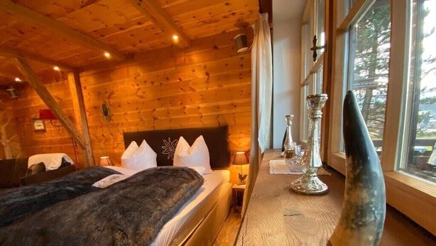 A wooden cabin room with a bed, a nightstand, a lamp, and a window with a view of trees. - Chalet "Alpine Time-Out in Swiss Pine Wood" with Mountain View in Inzell near Ruhpolding (Inzell)