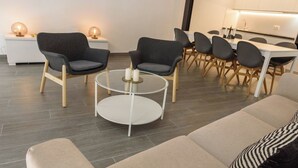 Ein modernes Wohnzimmer mit einem runden Couchtisch, zwei grauen Sesseln, einem beigen Sofa und einem Essbereich mit weißem Tisch und schwarzen Stühlen.