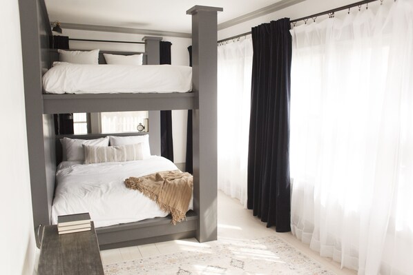 Habitación con literas de estructura gris, ropa de cama blanca y mesita de noche de madera.