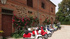 Une rangée de scooters anciens est garée devant un bâtiment en briques, doté d’arcs en plein cintre au niveau des fenêtres et d’une allée gravillonnée.