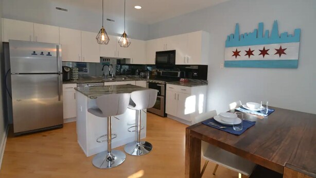 A modern kitchen with white cabinets, a stainless steel refrigerator, a microwave, and a sink. There are two white bar stools and a wooden dining table with plates and glasses. - Chic 2-Bedroom Townhouse with Rooftop, Free Parking, in Downtown Chicago! (Chicago)