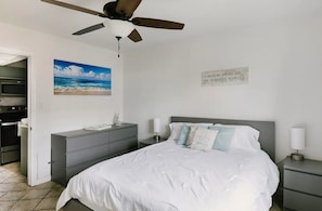 A bedroom with a bed, a ceiling fan, a painting of a beach, and a motivational quote on the wall.