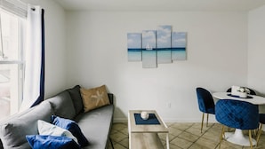 Ein modernes Wohnzimmer mit einem grauen Sofa, einem kleinen Tisch und einem blauen Sessel. Ein an der Wand hängendes Kunstwerk zeigt ein Segelboot auf dem Meer.