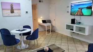 Ein Wohnzimmer mit Fernseher, einem kleinen Tisch, zwei blauen Stühlen und einem Bild von einer Ananas.