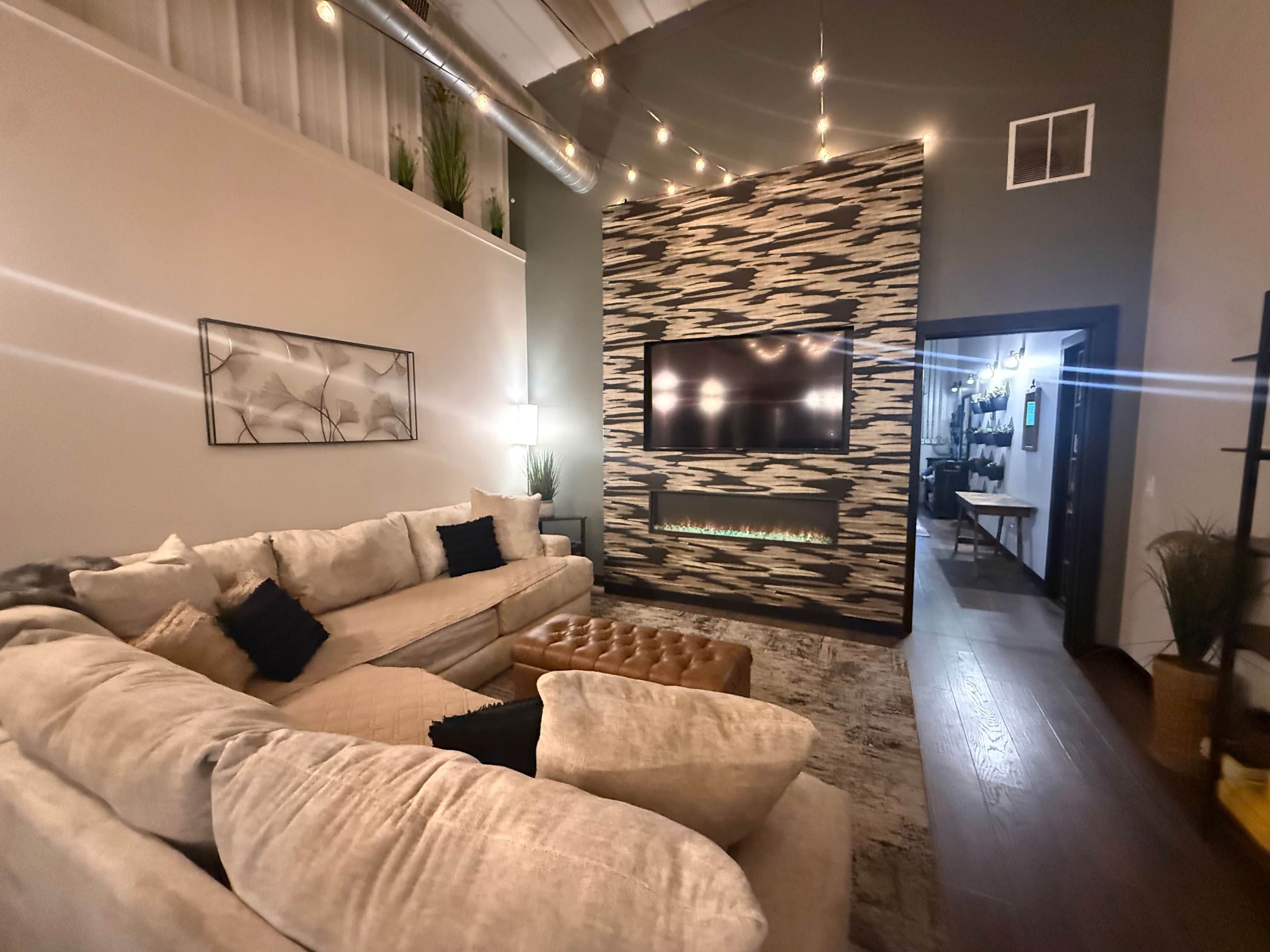 Un salon moderne avec un canapé d’angle, un téléviseur à écran plat fixé sur un mur texturé, une cheminée et un escalier menant à un niveau supérieur.