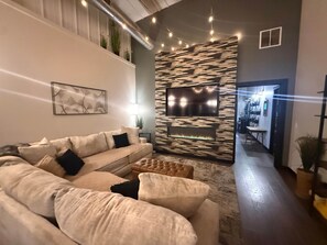 A modern living room with a sectional sofa, a flat-screen TV mounted on a textured wall, a fireplace, and a staircase leading to an upper level. - Feathered Nest at Fellows Lake w/ Indoor POOL! (Fair Grove)