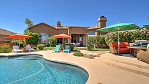 A swimming pool with lounge chairs and umbrellas, a patio area with outdoor furniture, and a house with a large window and a chimney. - Luxe Scottsdale Home w/ Pool & Mountain Views! (Scottsdale)