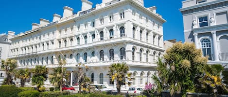 A large, historic building with multiple stories, ornate architectural details, and a well-maintained garden with palm trees.
